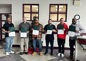 Students holding certificates stand in a row in front of a wall with two windows above their heads.