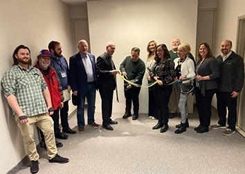 People stand in a row with a man holding a giant pair of scissors cutting two ribbons at the center.