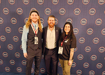 Three people stand together and smile in front of a backdrop with a circular logo and the letters "SVA" in the middle.