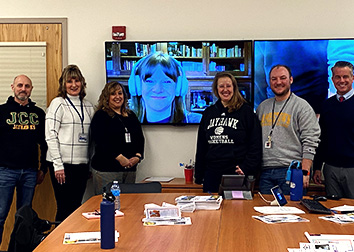 A group of people stand side-by-side with a monitor with a person's face on the wall in the center of the group.