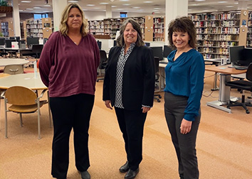 Three women stand together in an open area of a library.