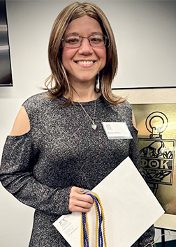 A woman stands holding an envelope and commencement cords in an indoor area with framed images on the walls.