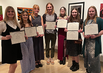 Seven students hold certificates while standing in front of a gallery wall with works of art hanging on it.