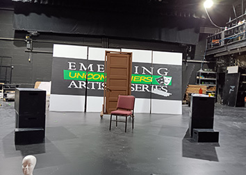 A model of a head, stacks of crate-sized cubes, a door on wheels and a chair form a set on stage in the Scharmann Theatre at SUNY JCC with a sign in the back that says "Uncommoners Emerging Artists Series."
