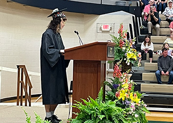 A woman in a cap and gown standing at a podium with people in bleachers around her.