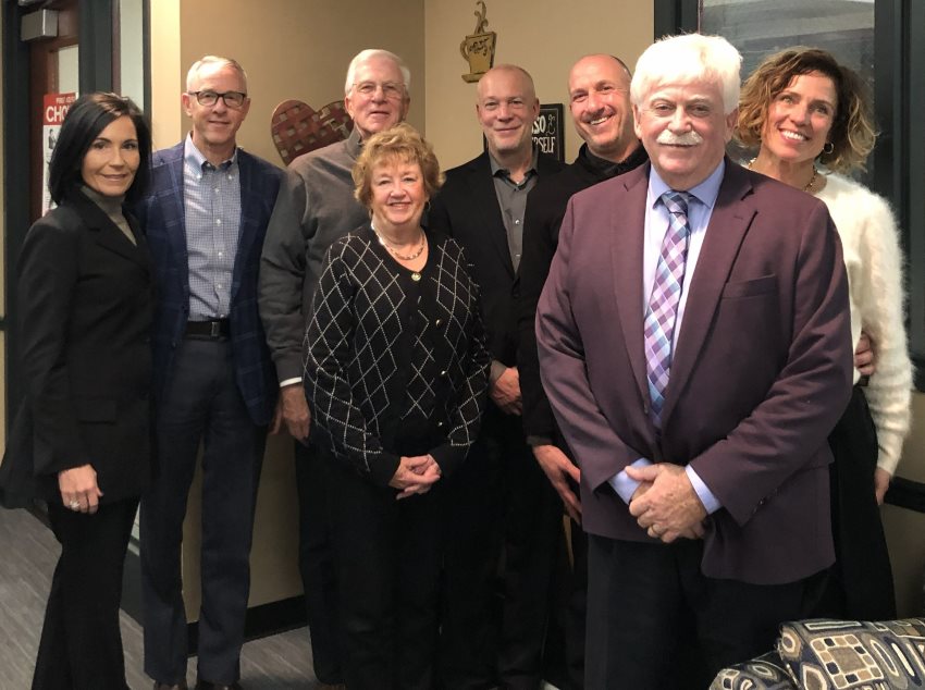 A group of eight adults, five men and three women, dressed in business attire, smile warmly while standing closely in a cafe setting.