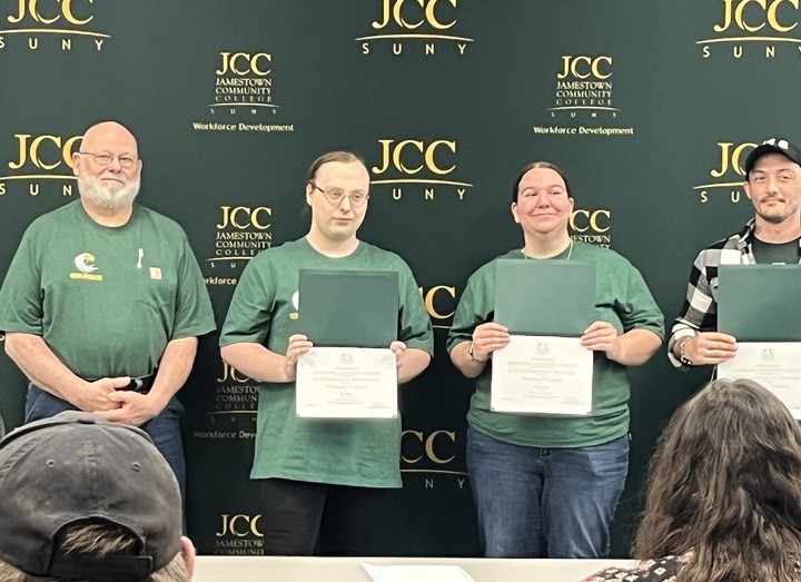 Four people stand in front of a "JCC SUNY Jamestown Community College" backdrop, holding certificates and smiling, conveying accomplishment.