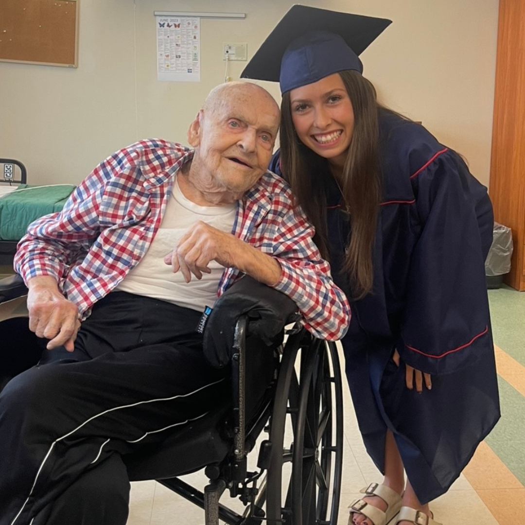 Woman in graduation cap and gown poses for a photo with an elderly man in a wheelchair.