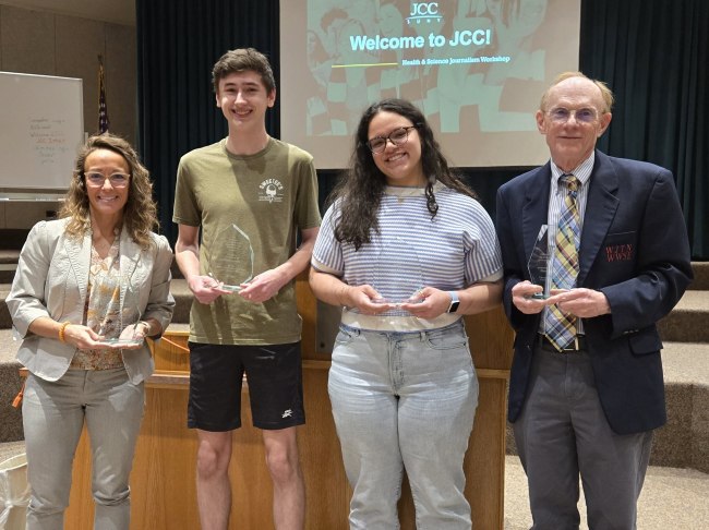 Four people stand together holding awards, smiling. Behind them, a screen reads "Welcome to JCC!" indicating a journalism workshop event.