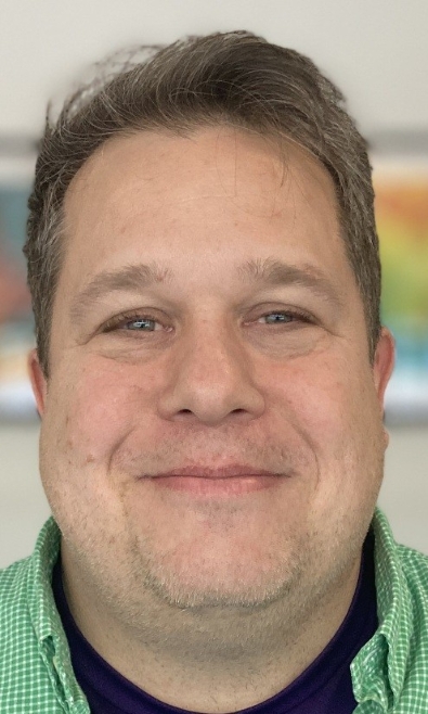 Close-up portrait of a man with short, graying brown hair, fair skin, and blue eyes, smiling. He is wearing a green and white checkered button-down shirt over a purple t-shirt.