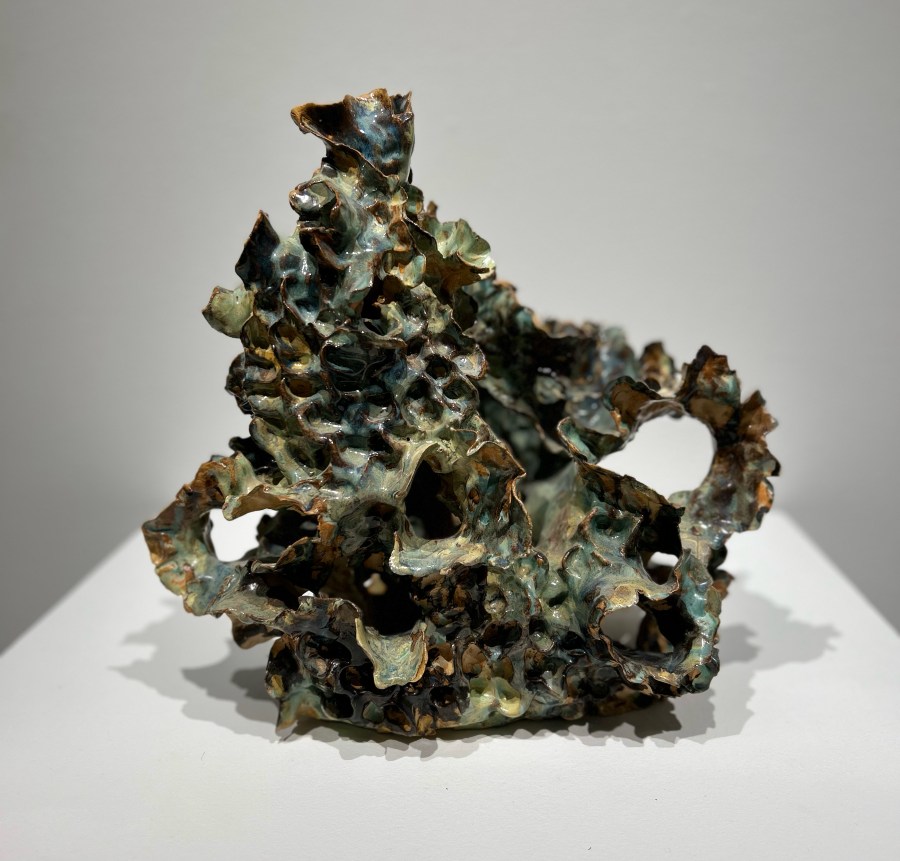 A rugged abstract bronze sculpture with jagged, porous surfaces and irregular openings stands on a white plinth, evoking ancient ruins and quiet mystery.