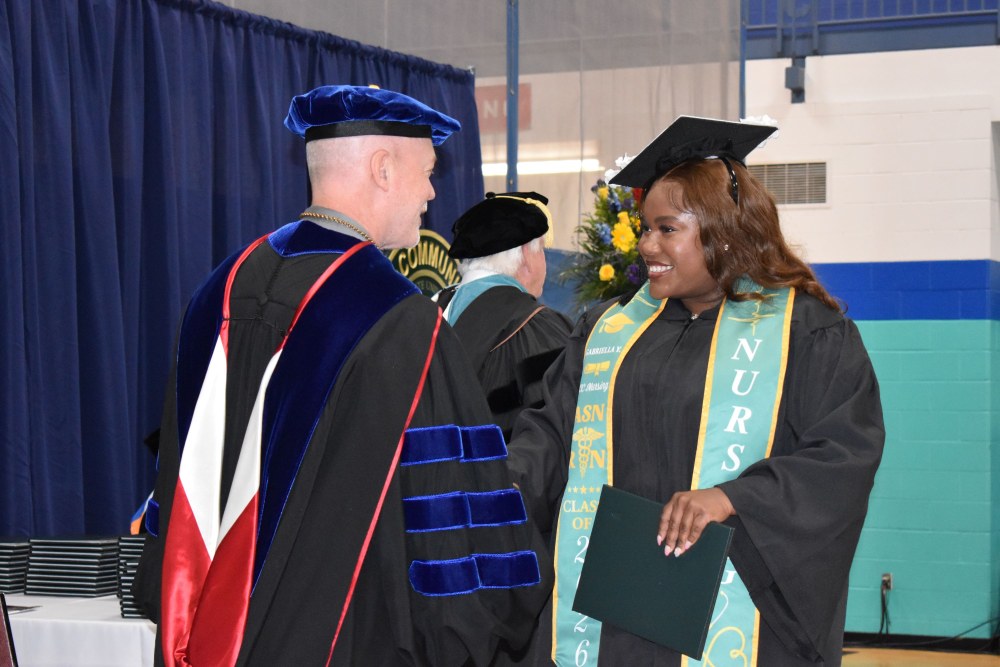 Graduation ceremony with a smiling graduate in cap and gown shaking hands with an official. The graduate wears a nursing stole and holds a diploma.