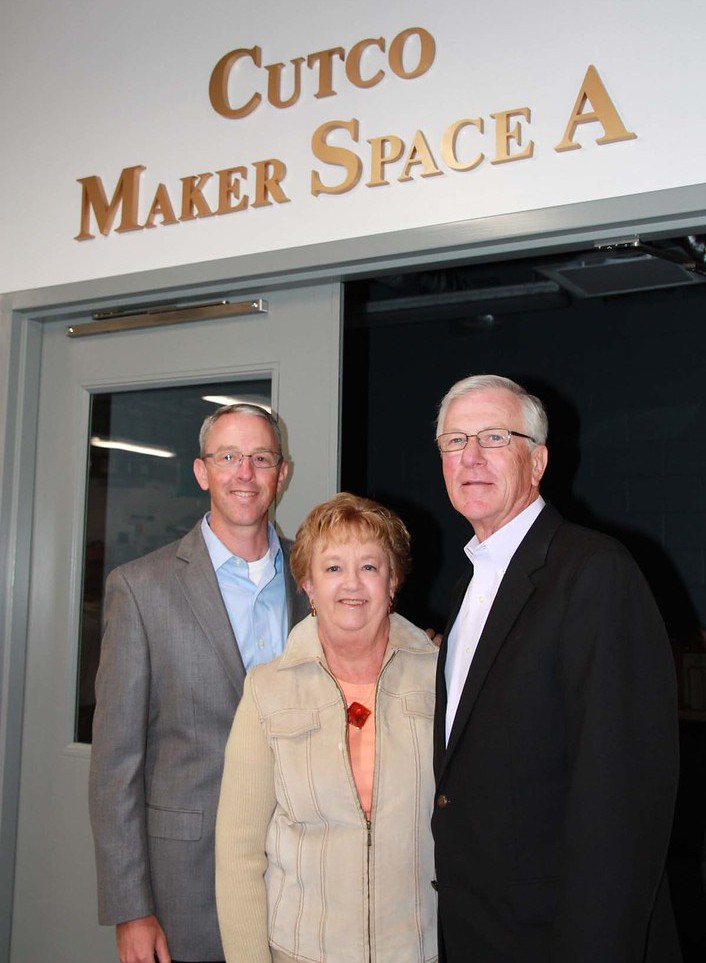 Three smiling adults pose beneath a “Cutco Maker Space” sign: two men in suits flank a woman in a beige jacket at an indoor entrance.