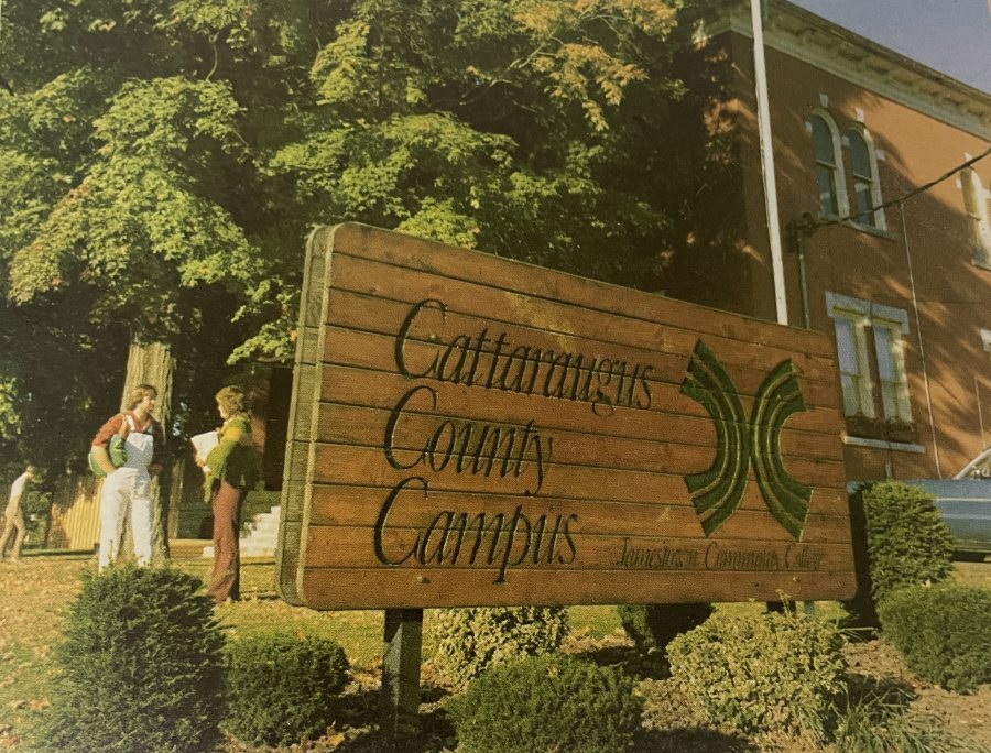 Wooden sign reading “Cattaraugus County Campus” stands amid manicured shrubs, trees, and students outside a historic brick building.