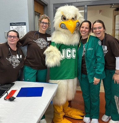 Healthcare staff smile beside a friendly JCC Jayhawk mascot in a green JCC shirt at an indoor event, with a decorated table in the foreground.