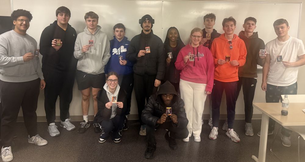 A diverse group of 12 young adults poses indoors, smiling and holding trading cards. They stand and kneel casually.