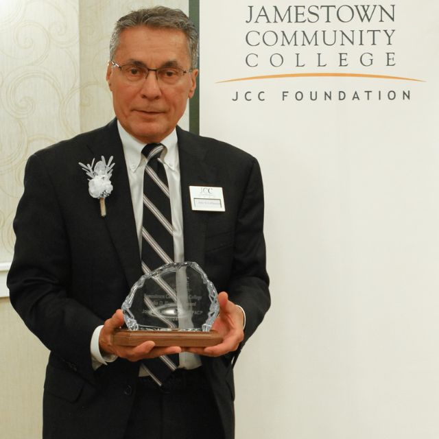 John M. LaMancuo holding his award with a SUNY JCC banner in the background.