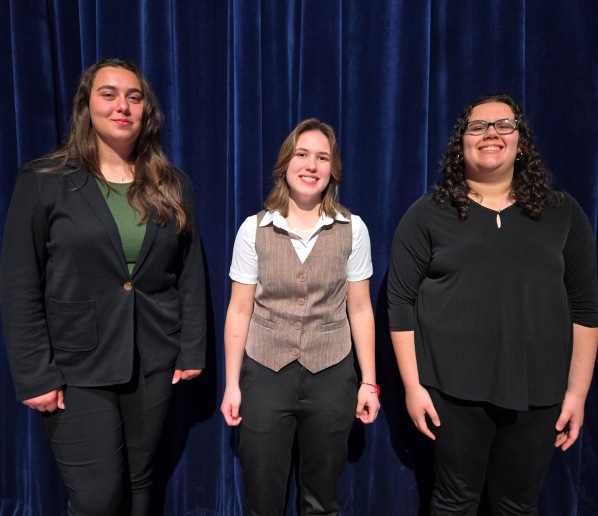 Three individuals stand in front of a dark blue curtain. The person on the left wears a black blazer over a green shirt and black pants. The person in the center wears a brown vest over a white shirt and black pants. The person on the right wears a black top and black pants.