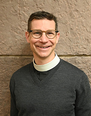 A man wearing a sweater and glasses stands near a wall.