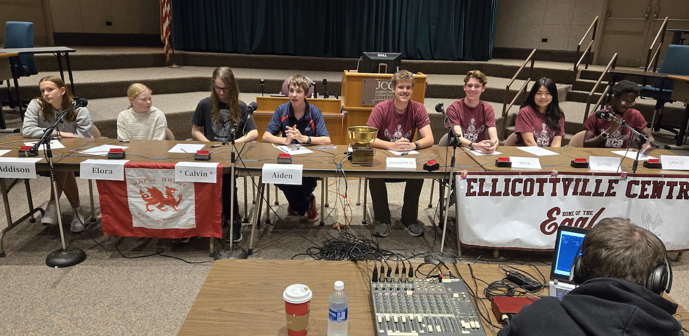 Eight students seated at long tables with microphones compete in a quiz competition.