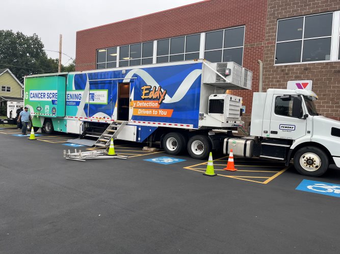 A large mobile screening truck with “Cancer Screening” and “Early Detection” signage is parked in a lot. Safety cones and handicapped spaces are visible.