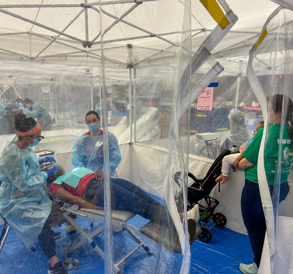 A patient receives dental care from two professionals in a medical tent while a woman holding a baby looks on.