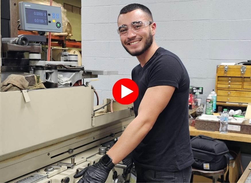 A person wearing safety glasses and gloves operates a machine in a workshop. They are smiling, conveying a positive and confident atmosphere. Overlayed by a red video play button.