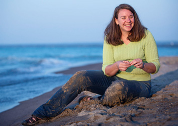 A woman sits by along the shore of a large body of water.