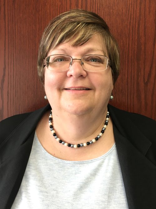 Close-up studio portrait of a woman wearing glasses, a black blazer over a light grey shirt, and a pearl and black beaded necklace against a brown wood-paneled background.