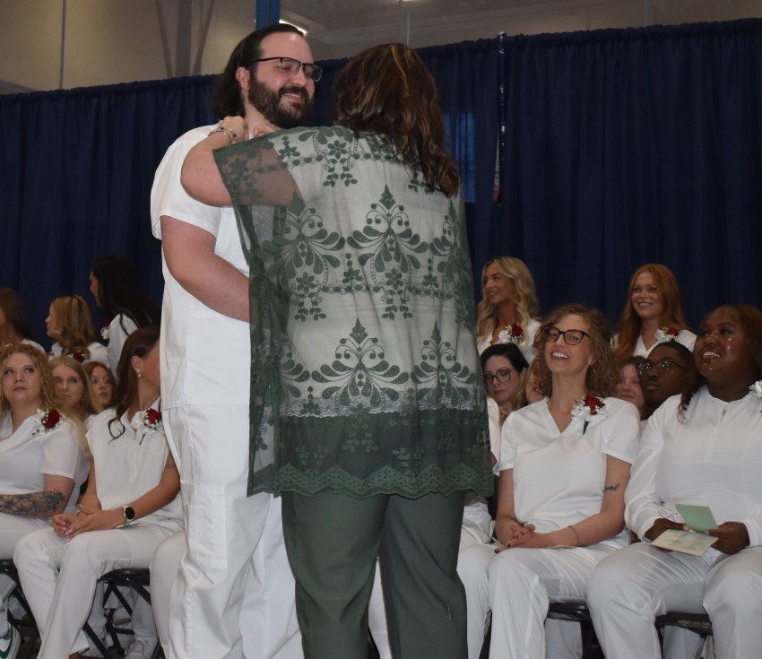 A man in scrubs is being presented with a green lace overlay by a woman. Several women in white scrubs are seated in the background.