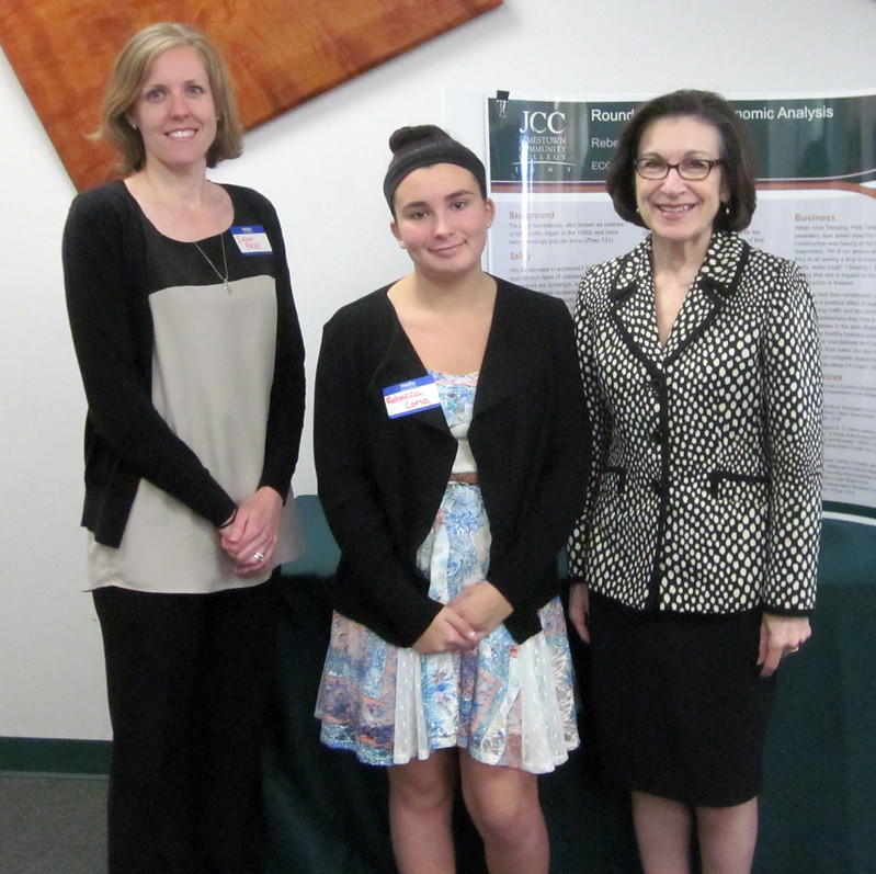 Three women pose smiling at an academic event, wearing name badges, with a research poster behind them; the mood is warm and professional.