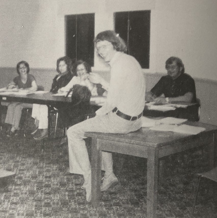 Black-and-white classroom photo of a smiling man in light clothing sitting on a desk, with students seated at tables behind him.