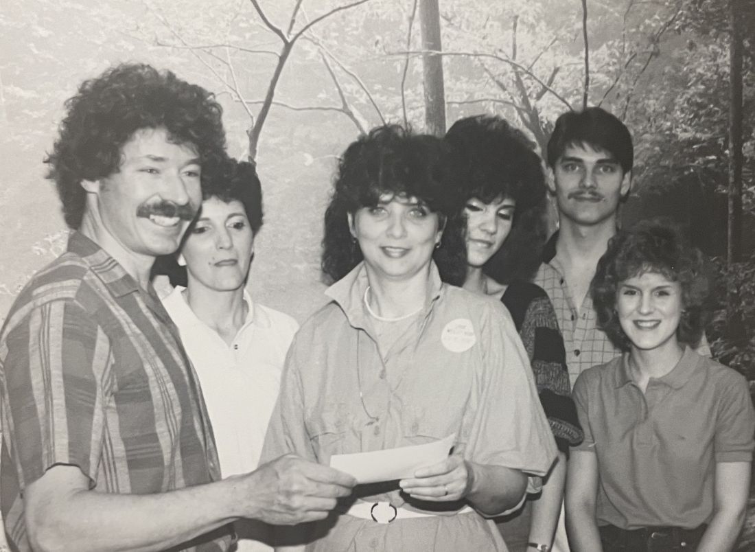 A group of six people stands in front of a painted backdrop of trees and branches. The person on the far left, a man with curly hair and a mustache, is wearing a striped shirt and is holding a piece of paper. Next to him is a woman with dark, short hair. In the center, a woman with dark, curly hair and a light-colored shirt is also holding a piece of paper, and she has a circular pin on her shirt. Behind her is another woman with dark, curly hair. To the right of the center woman stands a man with a mustache wearing a plaid shirt. On the far right is a woman with curly red hair wearing a collared shirt.