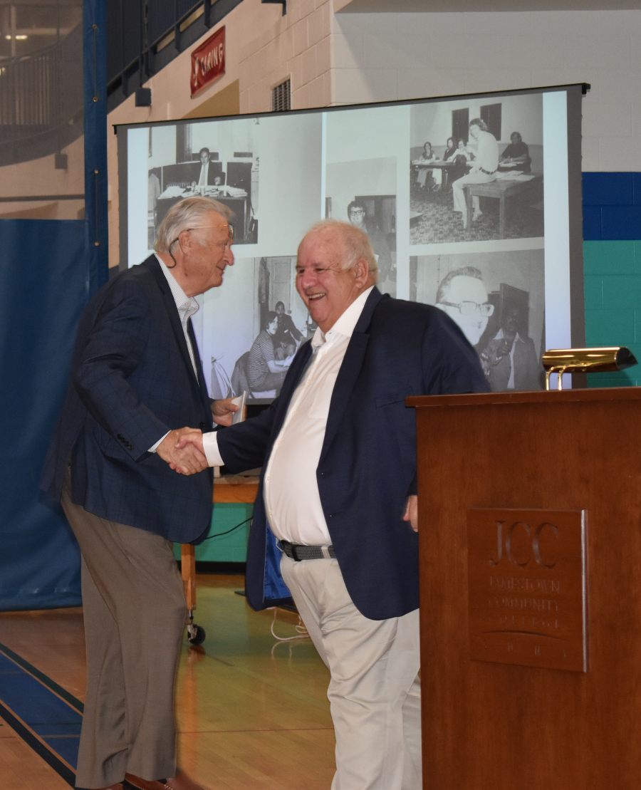 Two men in suits shake hands in front of a projector screen displaying historical black and white photos of people in rooms.