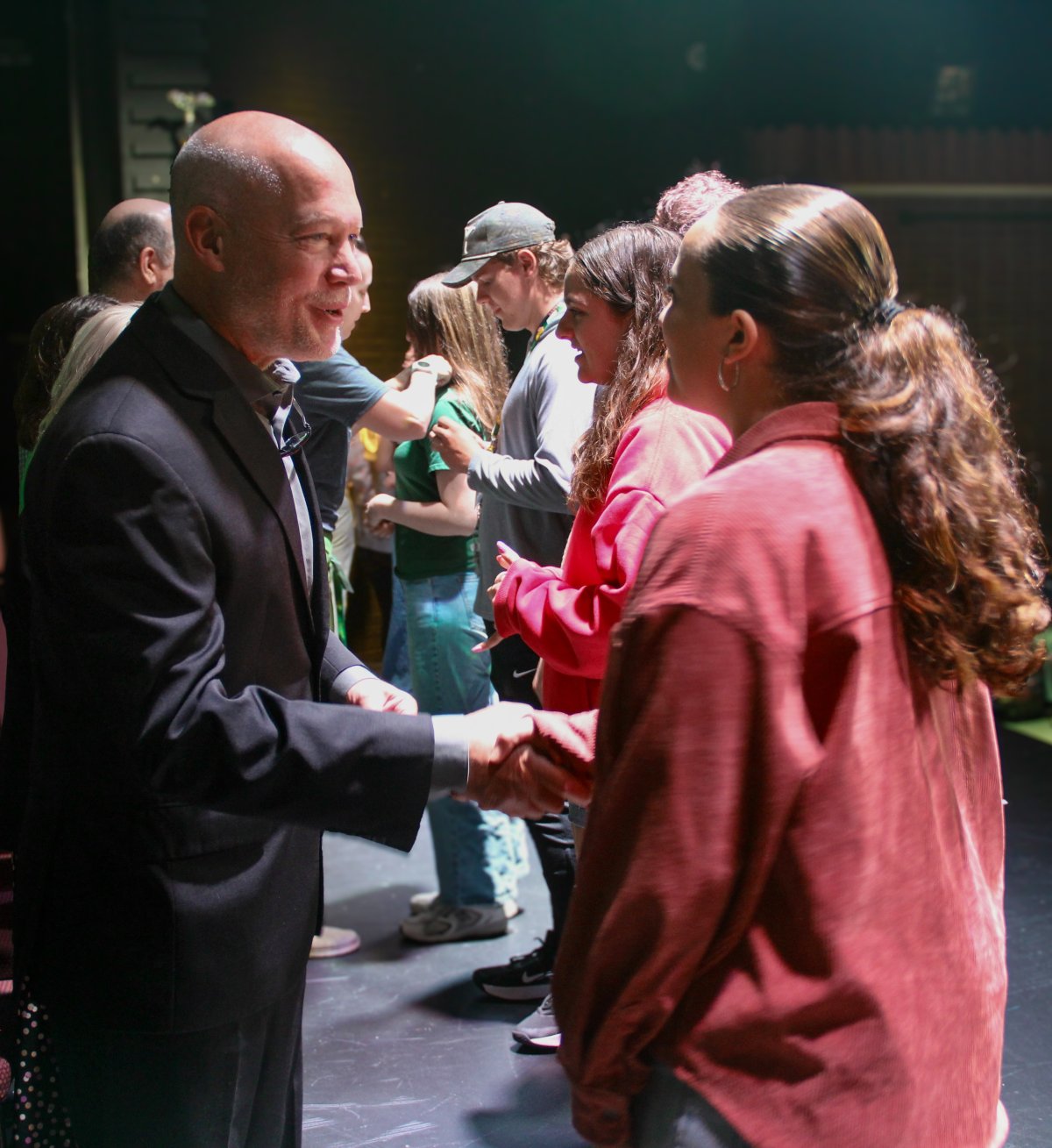 A bald man in a suit shakes hands with a young person wearing a red corduroy shirt while other young people stand in the background on a dark stage.