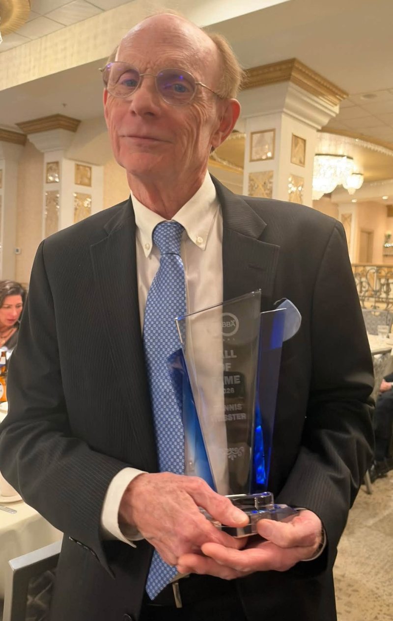 An elderly man in glasses and a dark suit holds a clear glass-and-blue award at a formal event, looking calm and proud in a warmly lit venue.
