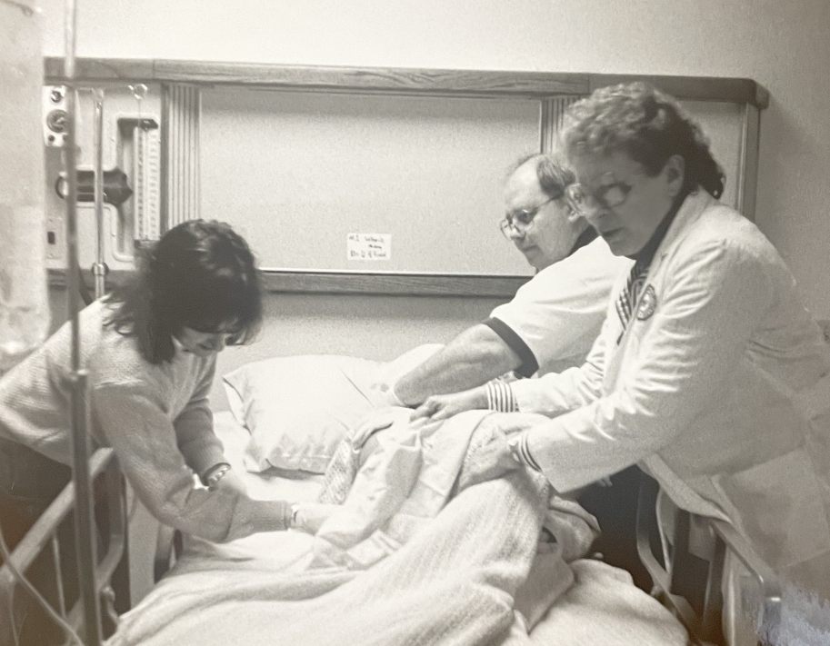Three individuals in a college nursing lab attend to a dummy patient in a hospital bed, with an IV stand visible on the left.