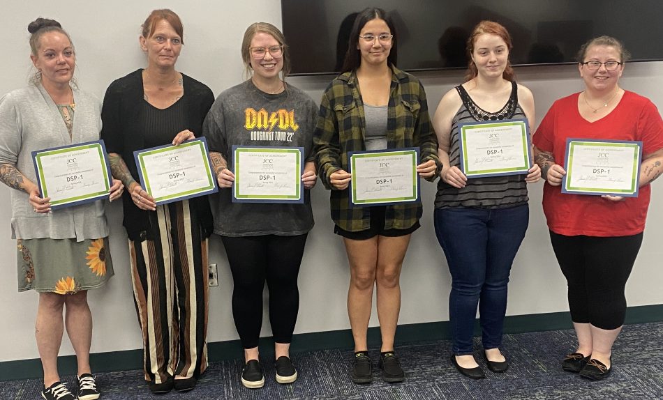 Six smiling women stand together indoors, each holding a framed certificate, celebrating achievement and recognition in a friendly group photo.