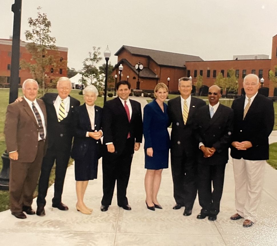 Eight diverse adults in formal attire pose outdoors on a campus walkway, smiling before a brick building and chapel in a friendly, professional scene.