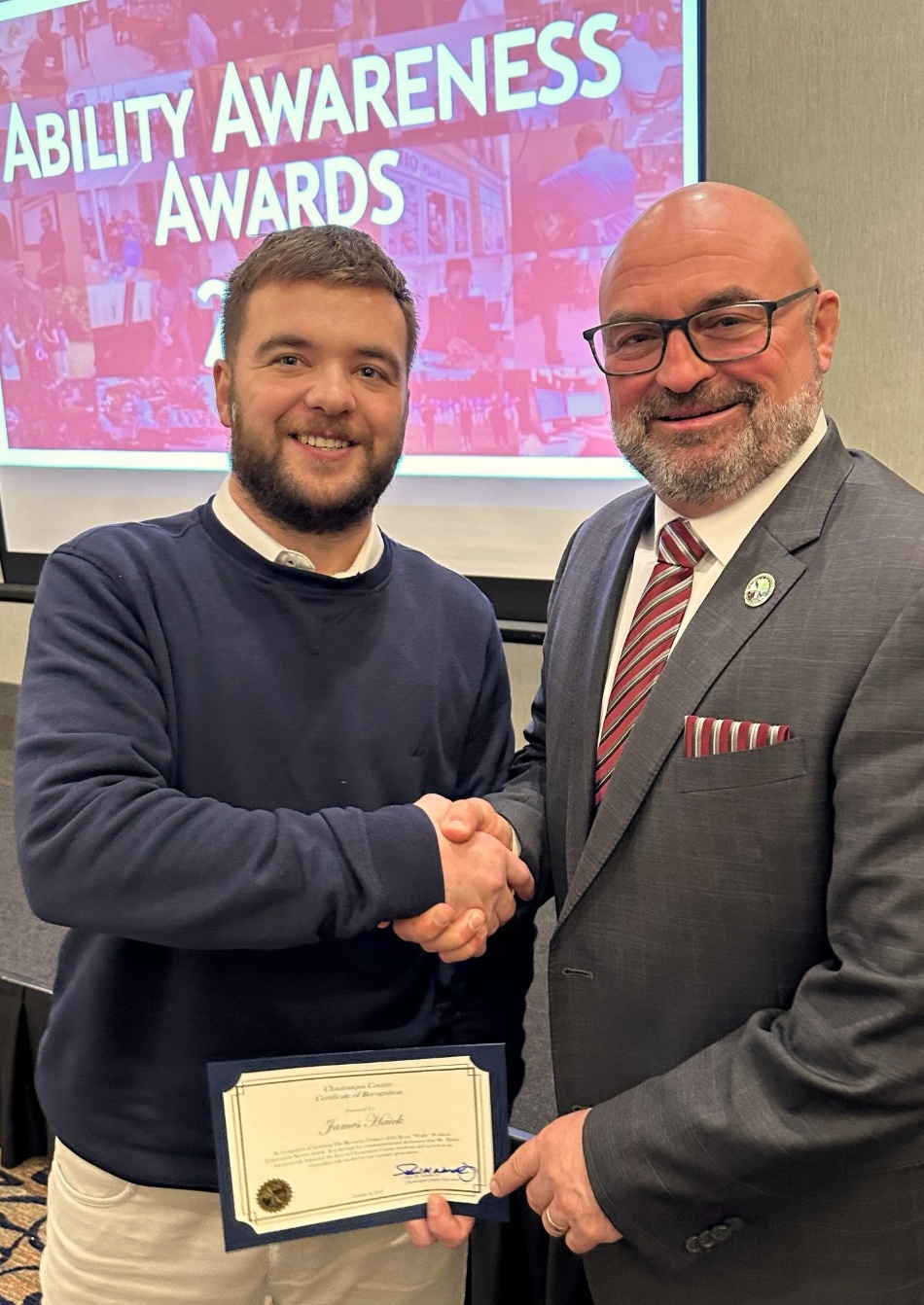 Two men smiling and shaking hands at the Ability Awareness Awards. One holds a certificate. Background shows a screen with the event name