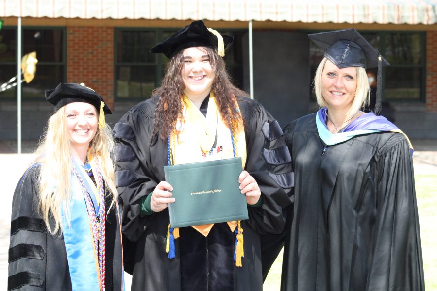 Three individuals in graduation gowns and caps smile, with the person in the center holding a green diploma cover.
