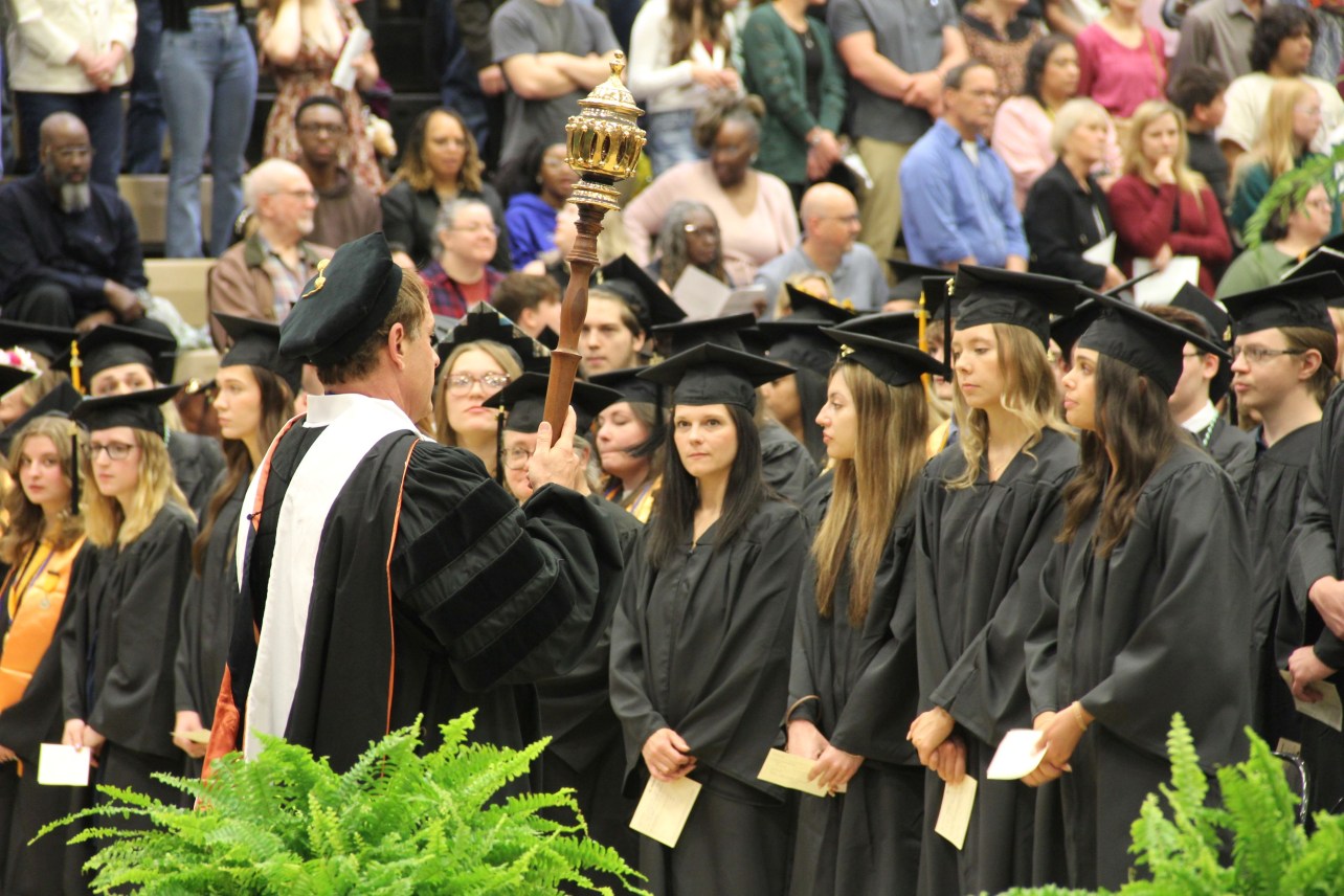 A professor in academic regalia holds a ceremonial mace, facing graduates in caps and gowns. An audience watches, with an atmosphere of formality and celebration.