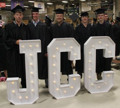 Five people wearing graduation caps and gowns stand behind giant JCC letters.
