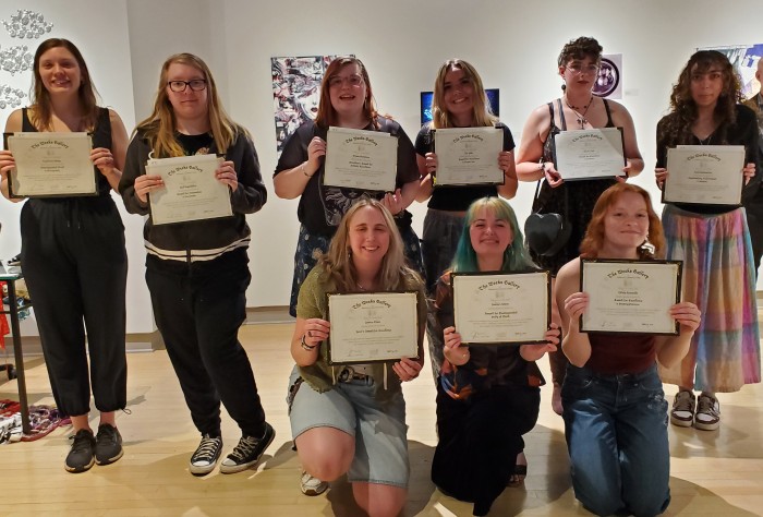 Nine people pose proudly indoors with certificates, smiling. The setting is a gallery with artwork in the background, conveying achievement and joy.