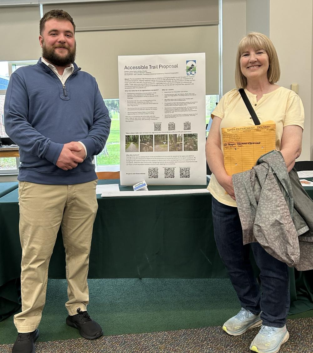 Two people stand smiling next to an "Accessible Trail Proposal" poster on an indoor table.