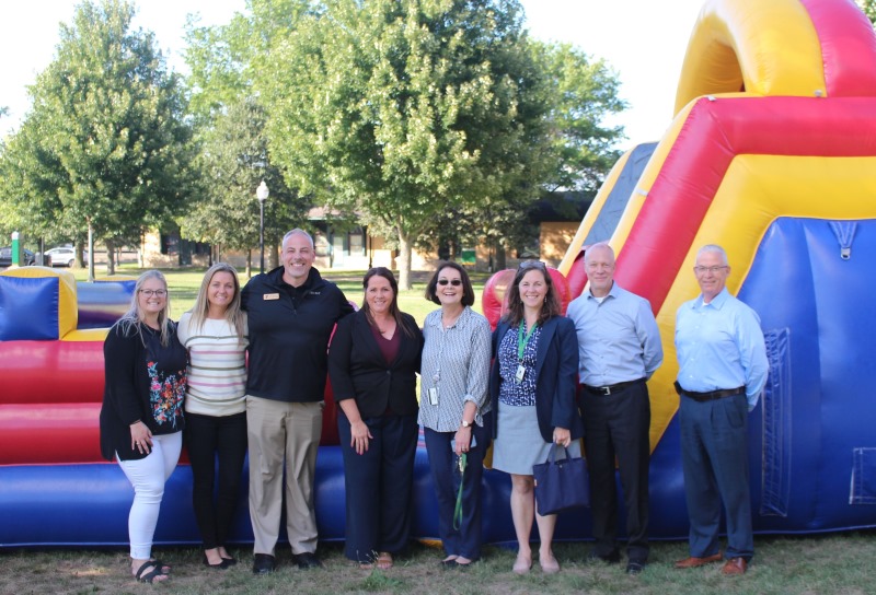 A group of eight adults stand smiling in front of a large inflatable obstacle course at an outdoor event. The group consists of men and women dressed in business casual attire, with trees and a grassy area visible in the background.