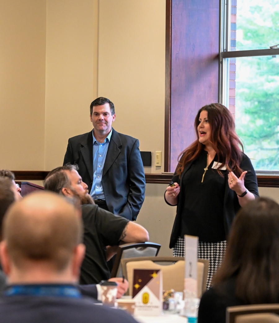 A man in a suit stands behind a woman in a black blazer speaking at a conference table in front of an audience.