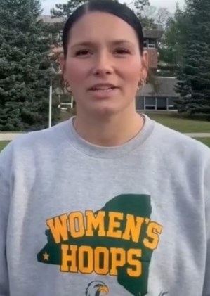 A person with short dark hair stands outdoors, wearing a gray sweatshirt displaying "Women's Hoops." Trees and a building can be seen in the background.