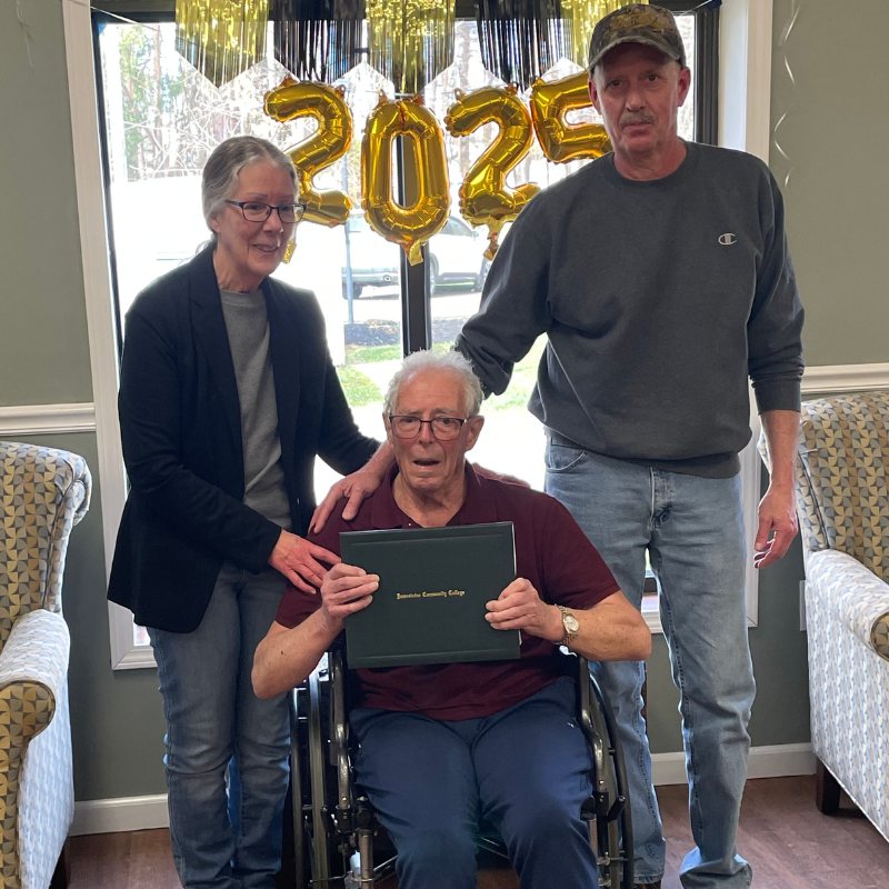 Two people stand on either side of a man a wheelchair who is holding a college diploma with a 2025 balloon hanging in the background.