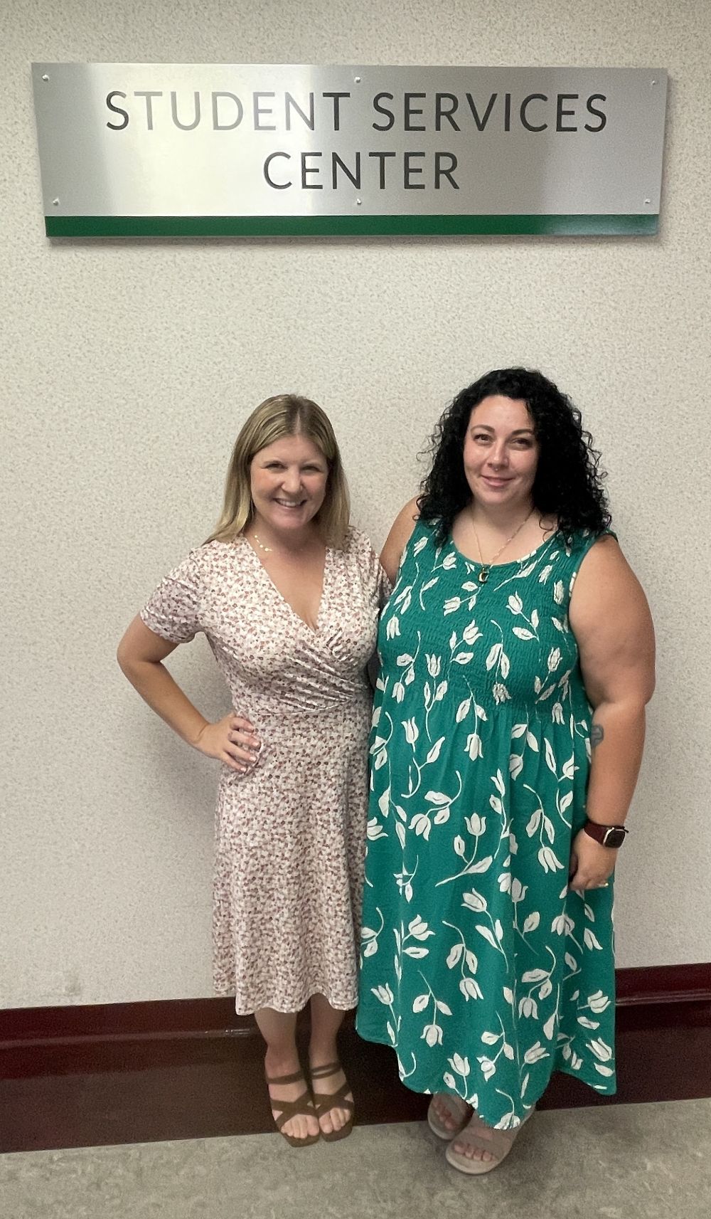 Two women stand side-by-side in front of a sign that reads "STUDENT SERVICES CENTER". The woman on the left wears a light-colored floral dress and brown sandals. The woman on the right wears a green floral dress with white flowers and light-colored sandals.